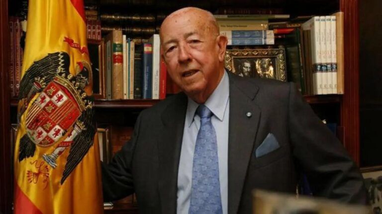 José Utrera Molina, ya anciano, posando en un despacho con estanterías de libros, vestido con traje oscuro y corbata azul, junto a una bandera de España desplegada, en una imagen de carácter institucional y alegórico.