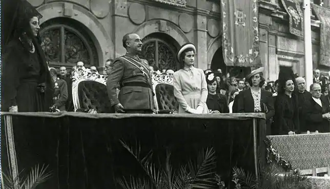 Fotografía histórica coloreada del acto de Franco en el Ayuntamiento de Sevilla (1953), con Franco de pie en la tribuna junto a varias autoridades y mujeres, ante la fachada del edificio consistorial y banderas oficiales de la época.