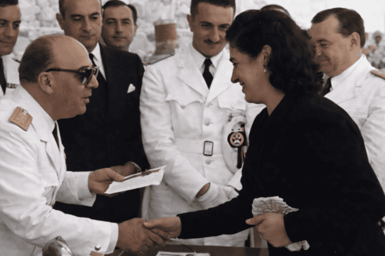 Fotografía histórica coloreada de 1952 en Pasajes de San Pedro: autoridades entregan documentación a una vecina durante el acto de inauguración de viviendas para pescadores, rodeados de representantes civiles y militares; escena de carácter social en un entorno portuario.