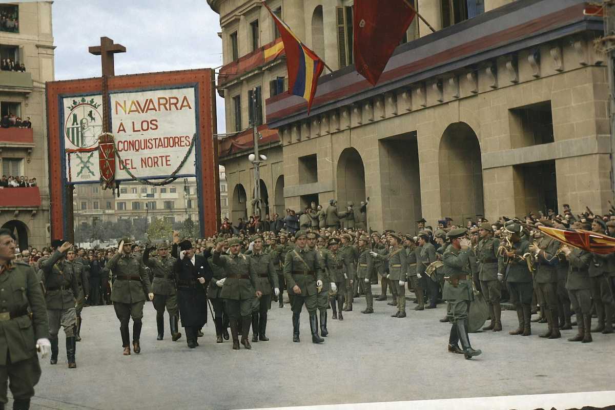 Fotografía histórica coloreada de un desfile militar en Pamplona durante la Guerra Civil española. Soldados con uniforme marchan por una plaza mientras una banda de música toca a la derecha. Al fondo se ve un gran arco con el lema “Navarra a los conquistadores del Norte”, banderas en los edificios y público observando desde balcones.