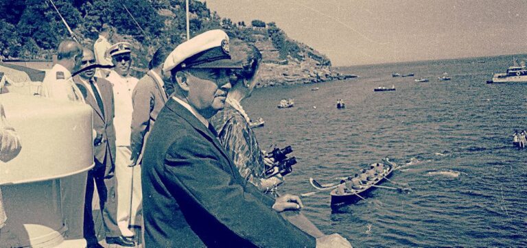 Fotografía histórica coloreada de Francisco Franco observando una exhibición naval desde una plataforma costera, acompañado de autoridades civiles y militares, con embarcaciones en el mar al fondo.