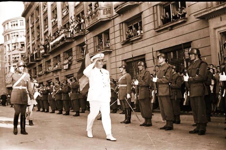 Desfile militar en una calle de Vitoria con un mando saludando a las tropas formadas, público en los balcones y bandera de España al fondo.