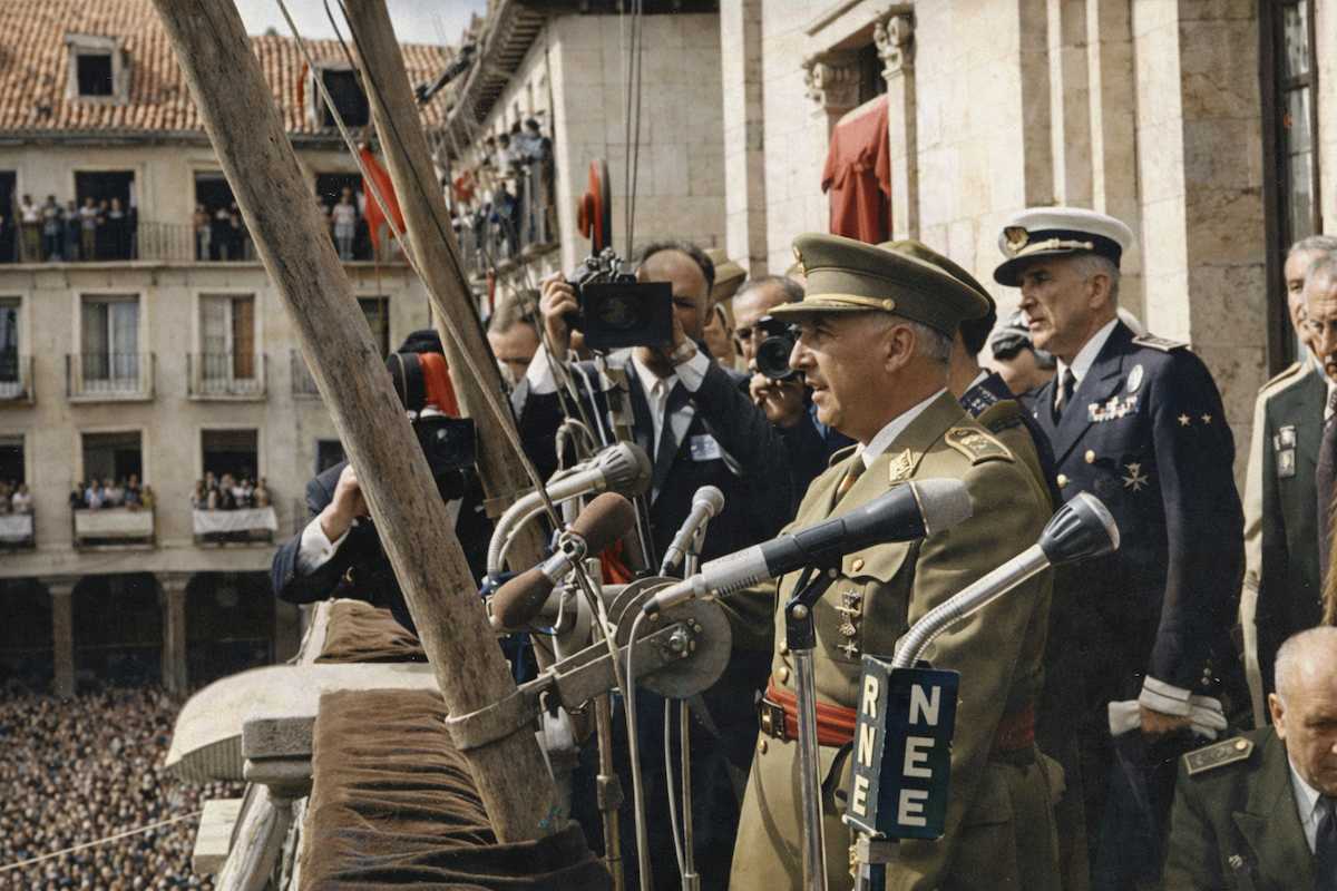 “Fotografía histórica coloreada de un discurso desde el balcón del Ayuntamiento de Ciudadela en 1960, con autoridades y micrófonos ante una multitud en la plaza.”
