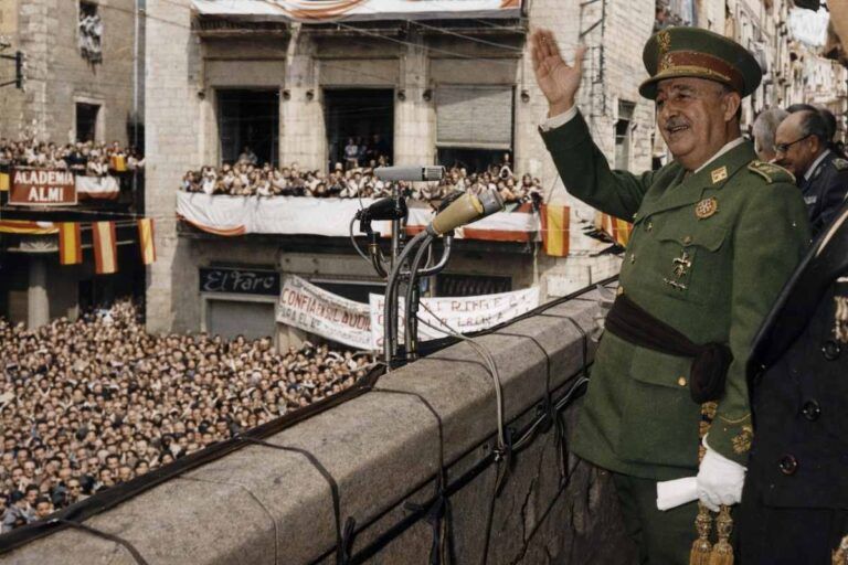 “Fotografía histórica coloreada de un discurso desde el balcón del Ayuntamiento de Gerona en 1960, con autoridades y una multitud congregada en la plaza.”