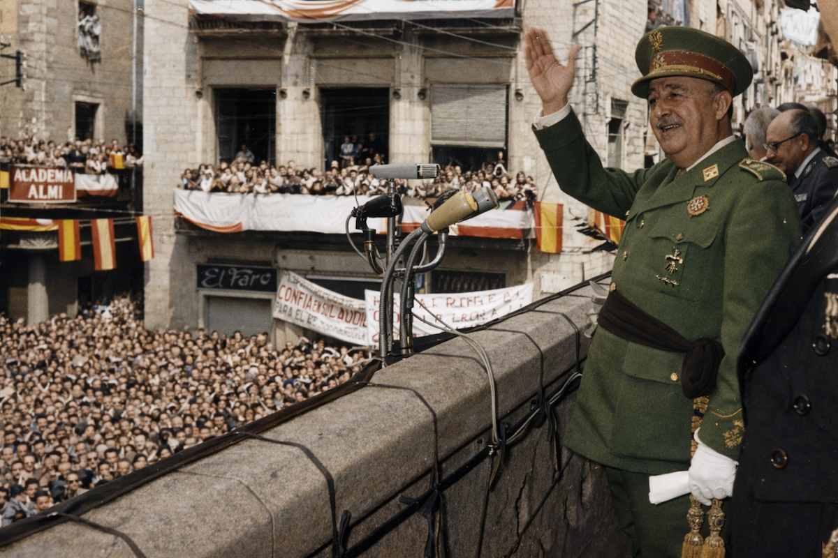 “Fotografía histórica coloreada de un discurso desde el balcón del Ayuntamiento de Gerona en 1960, con autoridades y una multitud congregada en la plaza.”