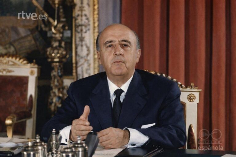 Fotografía histórica coloreada de un hombre sentado tras un escritorio durante una alocución televisada. Viste traje oscuro y corbata, gesticula con la mano mientras se dirige a la cámara. En primer plano se ven micrófonos y objetos de escritorio, y al fondo cortinas rojas y mobiliario de estilo clásico.