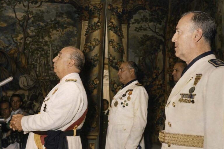 Francisco Franco, con uniforme blanco y condecoraciones, pronuncia un discurso durante las Jornadas Técnicas Sociales en Madrid (18 de julio de 1960), acompañado por altos mandos militares en un salón decorado con tapices históricos.