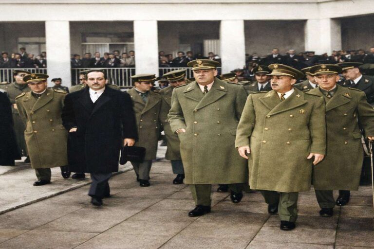 “Fotografía histórica coloreada de una inauguración oficial en 1960: varios altos mandos militares con abrigos y gorras caminan junto a un civil frente a un edificio con público al fondo.”