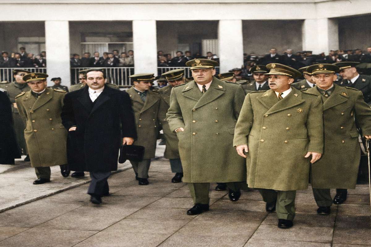 “Fotografía histórica coloreada de una inauguración oficial en 1960: varios altos mandos militares con abrigos y gorras caminan junto a un civil frente a un edificio con público al fondo.”