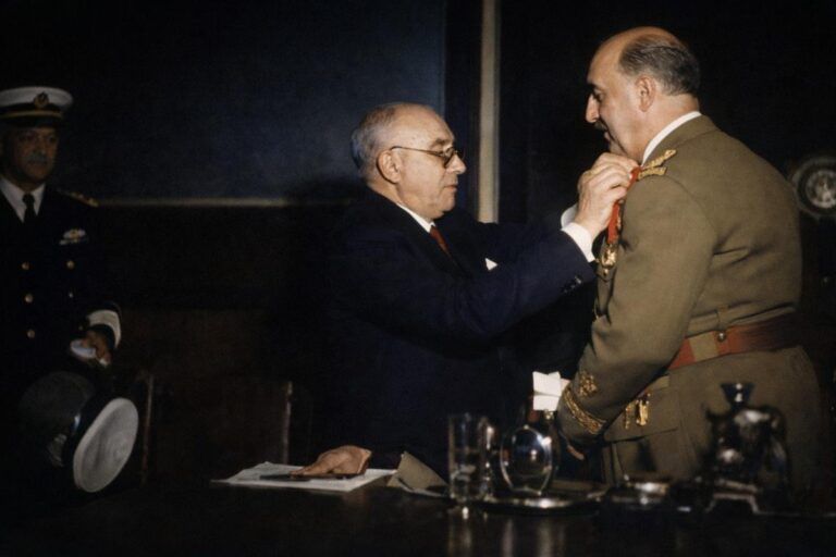 “Fotografía histórica coloreada del momento de la imposición de una medalla en un acto institucional en Mahón en 1960, con autoridades civiles y militares presentes.”