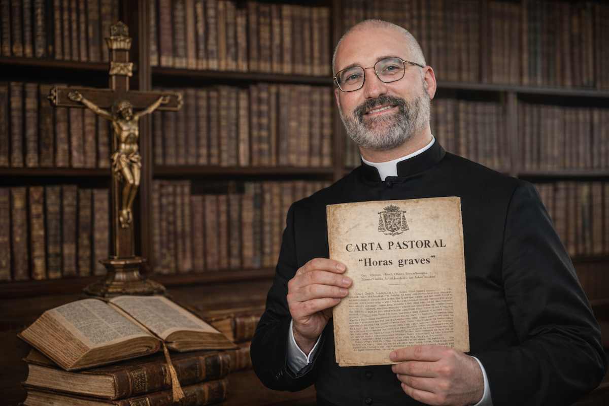 Sacerdote católico en una biblioteca antigua sosteniendo la carta pastoral «Horas graves», con un crucifijo y libros antiguos al fondo, en una escena alegórica.