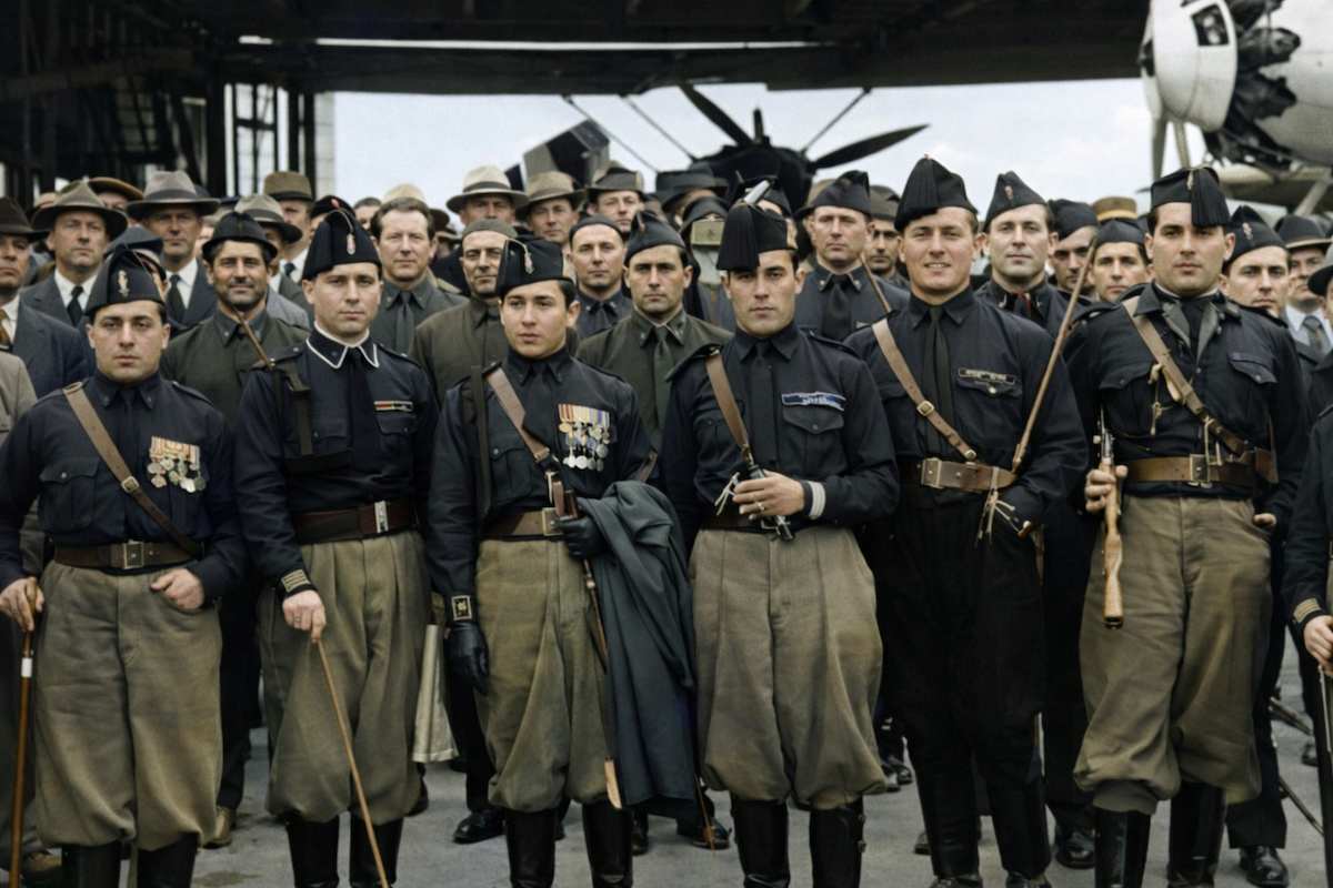 Fotografía histórica coloreada de un grupo de soldados italianos “camisas negras” posando en una pista aérea. Visten camisas negras, pantalones caqui y gorras militares; algunos portan fusiles y medallas en el pecho. Detrás de ellos se ven civiles y otros militares, así como un avión de la época en el hangar.