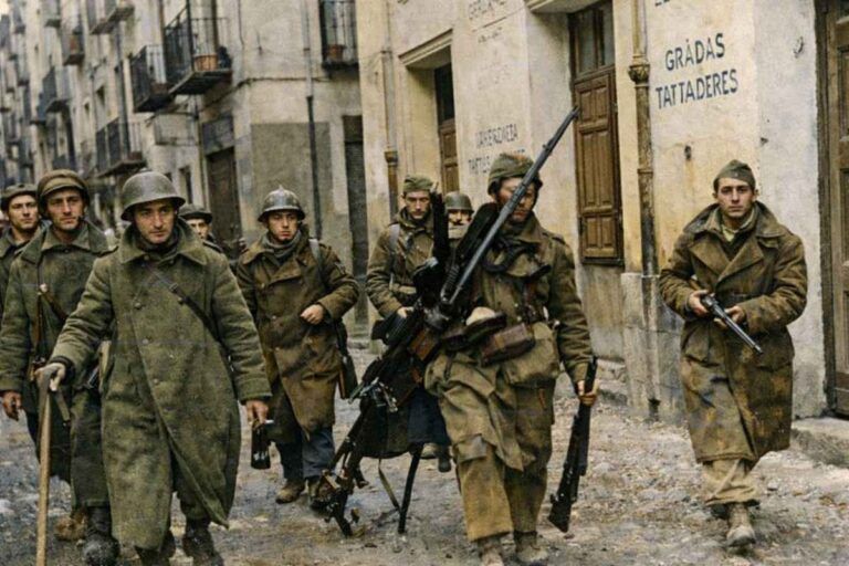 Fotografía histórica coloreada de soldados avanzando por una calle estrecha y dañada durante la Guerra Civil española. Visten abrigos y cascos de invierno y portan fusiles y una ametralladora ligera. Al fondo se ven edificios con fachadas deterioradas, lo que refleja un entorno urbano afectado por los combates.