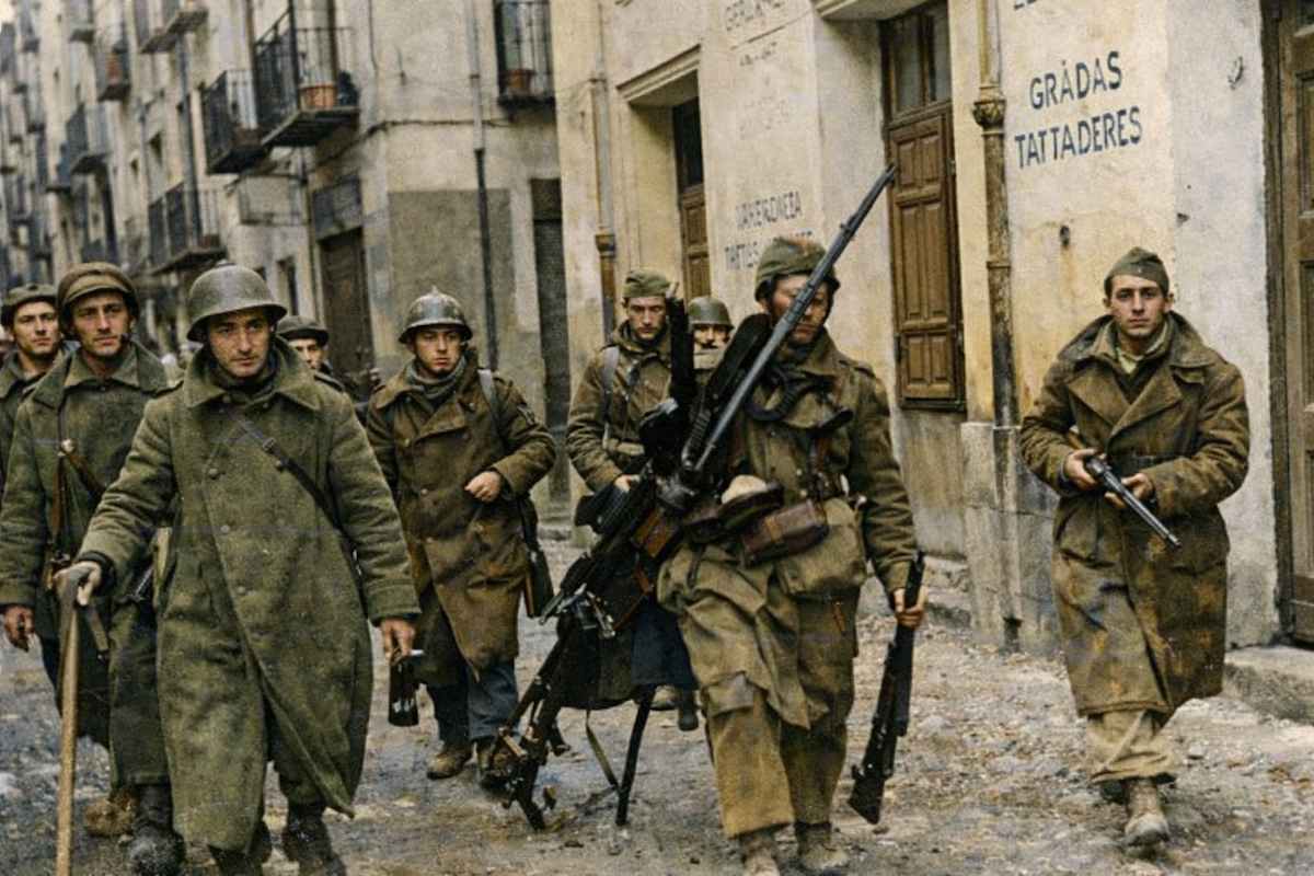 Fotografía histórica coloreada de soldados avanzando por una calle estrecha y dañada durante la Guerra Civil española. Visten abrigos y cascos de invierno y portan fusiles y una ametralladora ligera. Al fondo se ven edificios con fachadas deterioradas, lo que refleja un entorno urbano afectado por los combates.