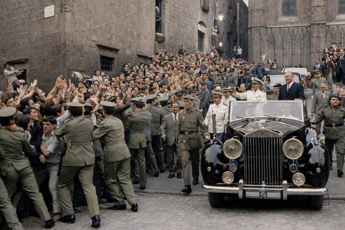 “Fotografía histórica coloreada de una visita institucional a la Ciudad Universitaria de Pedralbes en 1960, con un coche oficial rodeado de autoridades, fuerzas de seguridad y público.”