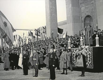 Acto oficial en un monumento con autoridades civiles y militares; un uniformado con traje verde militar destaca entre banderas y público reunido.
