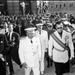 Franco en Vitoria, en su entrada al nuevo edificio, flanqueado por el gobernador civil de la provincia, Luis Martín-Ballestero Costea (a su izquierda), y por el alcalde de Vitoria, Gonzalo Lacalle Leloup (a su derecha). Tras ellos, al fondo, observamos al cámara grabando para el Noticiario Cinematográfico Español (NO-DO).
