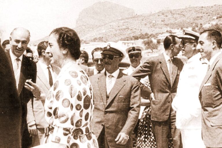 Francisco Franco conversando con autoridades civiles y militares durante una visita oficial en España en 1958, en un entorno costero acompañado por mandos navales y representantes locales.