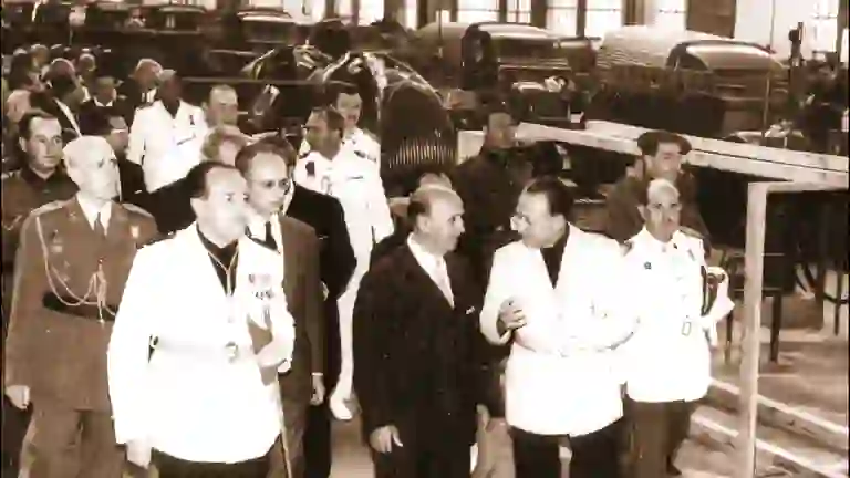 Francisco Franco visitando una fábrica industrial en España en 1958, acompañado por autoridades civiles y militares mientras recorren las instalaciones con maquinaria y vehículos industriales al fondo.