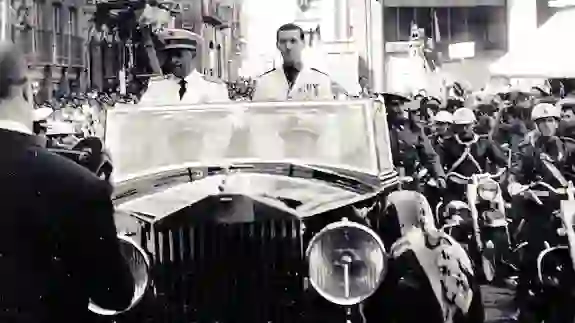 Fotografía histórica coloreada de Francisco Franco en un automóvil descapotable durante un acto público en una calle abarrotada, escoltado por motoristas y autoridades, con banderas españolas entre la multitud.