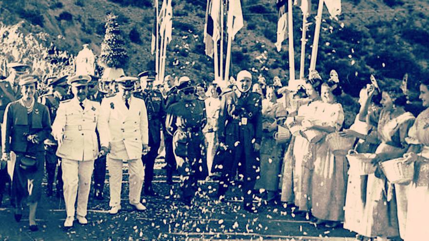 Fotografía histórica coloreada de Francisco Franco caminando junto a autoridades militares y civiles durante un acto oficial, mientras mujeres con trajes tradicionales lanzan pétalos de flores a su paso en una ceremonia pública en España.