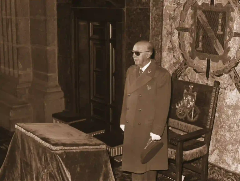 Francisco Franco en un acto institucional en 1958 durante la conmemoración fundacional de la Falange, de pie junto a una mesa ceremonial con el escudo de España y un tapiz con el emblema falangista al fondo.