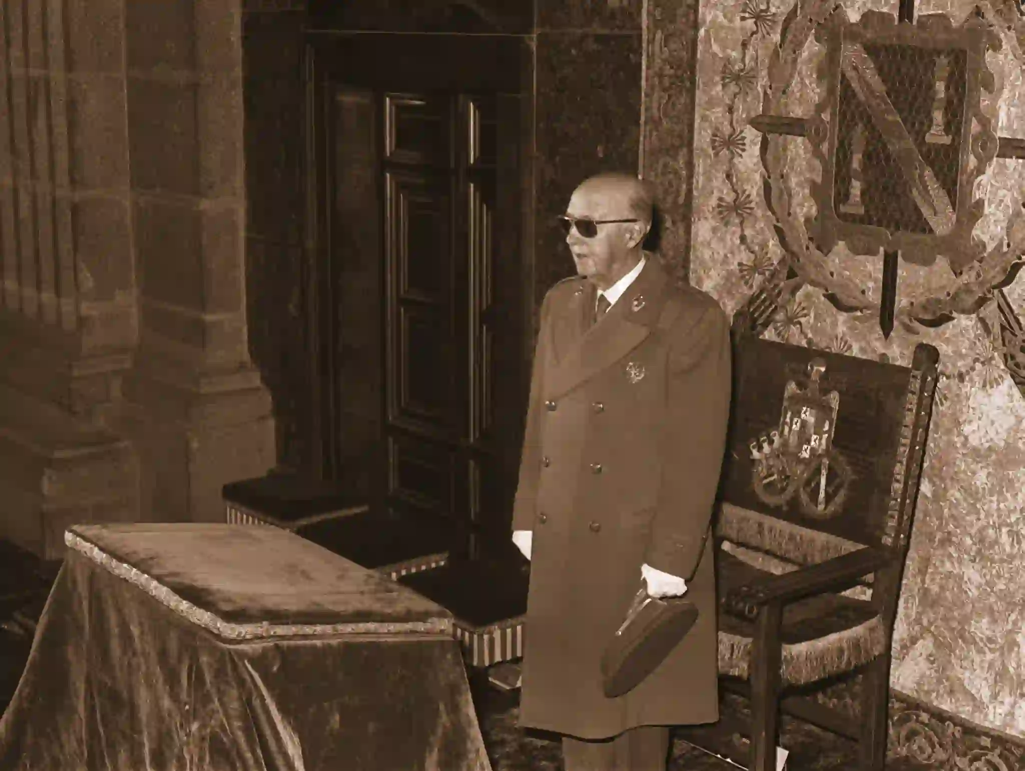 Francisco Franco en un acto institucional en 1958 durante la conmemoración fundacional de la Falange, de pie junto a una mesa ceremonial con el escudo de España y un tapiz con el emblema falangista al fondo.