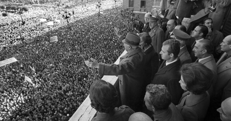 Franco saluda desde un balcón a una multitud congregada en la Plaza de Oriente durante un acto público, rodeado de autoridades y ante una gran concentración de personas.