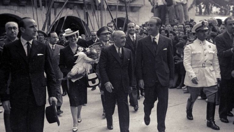 Francisco Franco caminando entre autoridades civiles y militares durante una visita oficial en Guipúzcoa en 1961, rodeado de público y acompañado por una mujer con un ramo de flores.