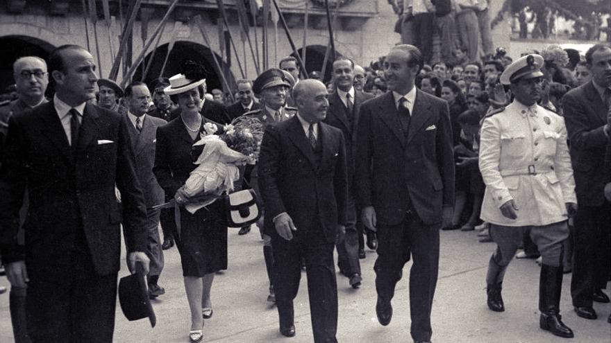 Francisco Franco caminando entre autoridades civiles y militares durante una visita oficial en Guipúzcoa en 1961, rodeado de público y acompañado por una mujer con un ramo de flores.