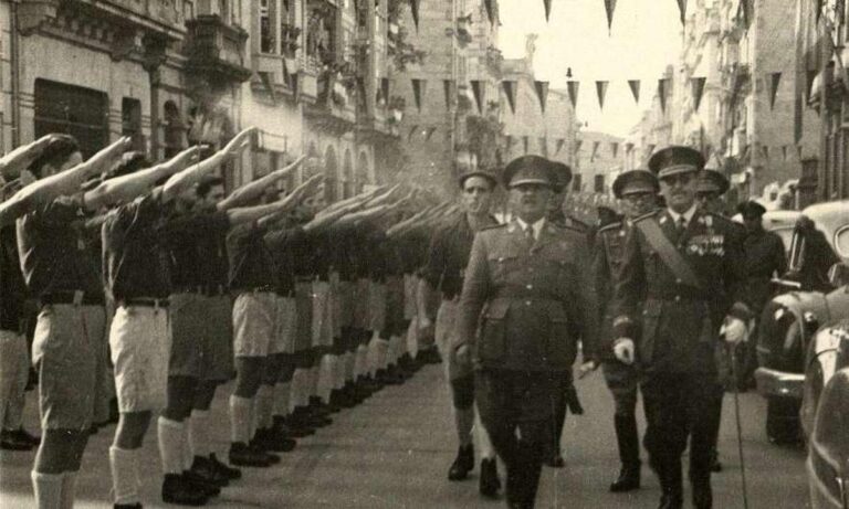 Desfile de recepción a Francisco Franco durante su visita a Ourense en 1961, con formación de jóvenes saludando en una calle engalanada con banderas mientras el jefe del Estado avanza acompañado por autoridades militares.