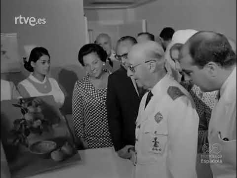 Primer plano de una escena institucional en blanco y negro donde Francisco Franco, vestido con uniforme militar, observa atentamente una exposición o maqueta sobre una mesa, acompañado por varias personas —hombres con traje y una mujer— en un entorno interior oficial.