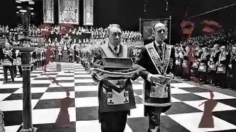 Ceremonia masónica en un templo con suelo ajedrezado, donde varios hombres vestidos con delantales y ornamentos rituales avanzan en formación mientras otros miembros observan desde los laterales.