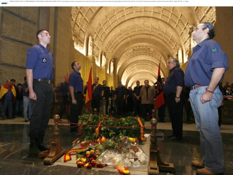 Acto conmemorativo en un edificio histórico con varias personas uniformadas y asistentes rodeando una corona de flores con cintas de colores, en un ambiente solemne y ceremonial bajo una bóveda arquitectónica.