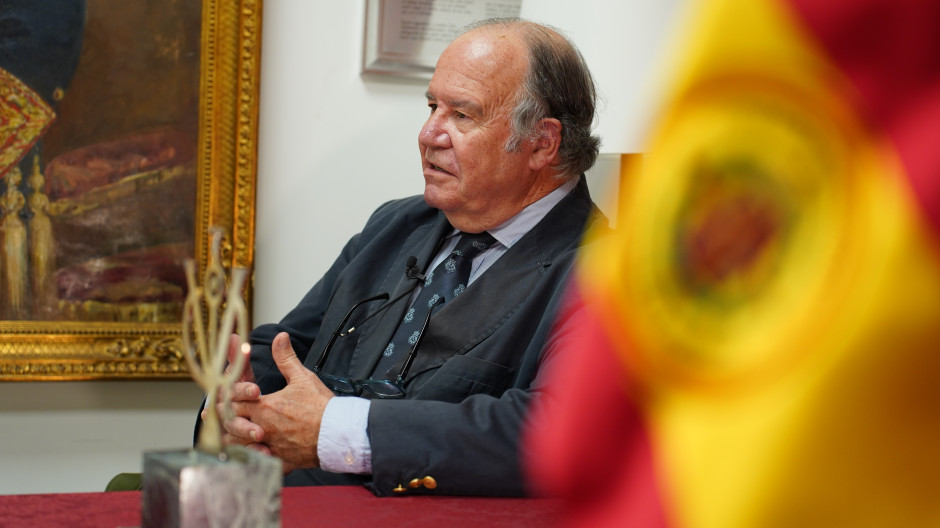 Hombre mayor con traje oscuro sentado en una mesa, hablando en una entrevista, con un cuadro enmarcado al fondo y una bandera de España desenfocada en primer plano.