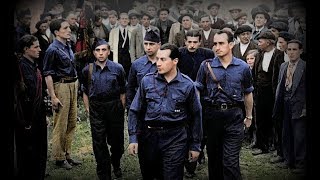Grupo de hombres uniformados caminando con determinación entre una multitud de obreros, en un entorno industrial, reflejando un ambiente de movilización social y tensión histórica.