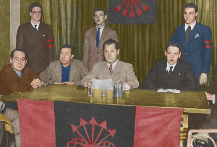 Fotografía en color de los años 30: José Antonio Primo de Rivera sentado en el centro de una mesa con varios hombres a su alrededor, algunos de pie y otros sentados, en un entorno interior con cortinas al fondo y una bandera con el símbolo del yugo y las flechas visible detrás y sobre la mesa.