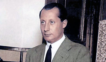 Retrato de José Antonio Primo de Rivera en primer plano, vestido con traje claro y corbata oscura, mirando ligeramente hacia un lado en un entorno interior sobrio.