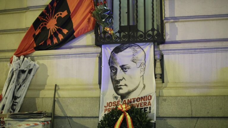 Homenaje con bandera y símbolo del yugo y las flechas junto a un cartel con retrato de José Antonio Primo de Rivera en una fachada, acompañado de flores con los colores de España.