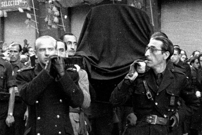 Fotografía histórica en blanco y negro de varios hombres transportando a hombros un ataúd cubierto con una tela, rodeados de una multitud en una calle durante un cortejo fúnebre.