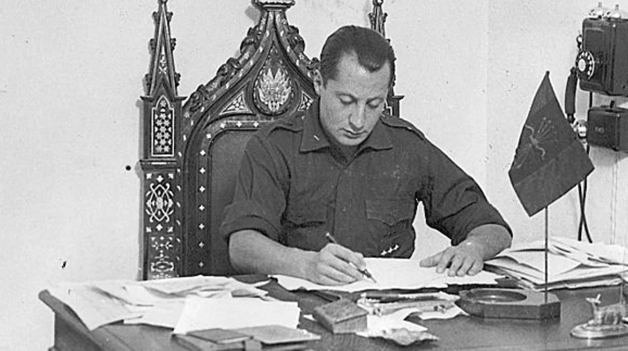Fotografía histórica en blanco y negro de un hombre sentado en un despacho, escribiendo sobre documentos en una mesa, con una silla ornamentada detrás y una pequeña bandera sobre el escritorio.
