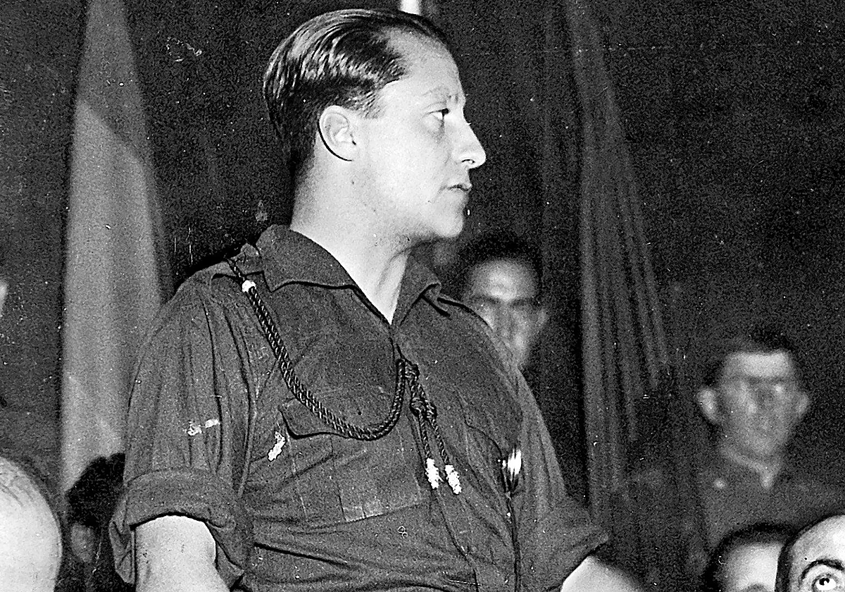 Fotografía histórica en blanco y negro de un hombre de perfil, vestido con uniforme, durante un acto público en interior, con otras personas desenfocadas al fondo.